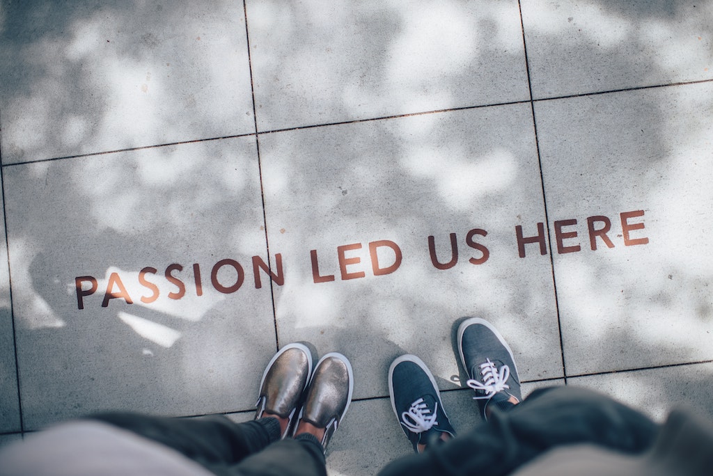 Two people stand on a sidewalk with "Passion led us here" painted on the ground below their feet.